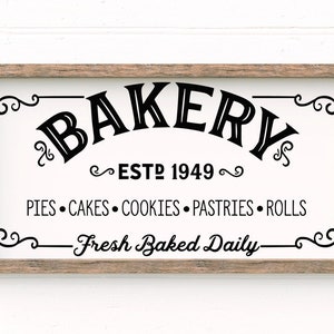 Pode incluir: Uma placa branca com texto preto que diz "BAKERY ESTD 1949" e "PIES CAKES COOKIES PASTRIES ROLLS -Fresh Baked Daily- ". A placa é emoldurada em uma moldura de madeira marrom.