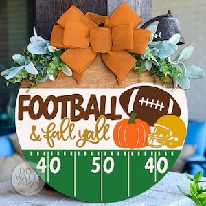 以下が含まれることがあります： 「FOOTBALL & fall y'all」の文字が入った丸い木製の看板。フットボール、カボチャ、ヘルメットが描かれています。オレンジ色のリボンと緑で装飾され、フットボールフィールドのデザインになっています。