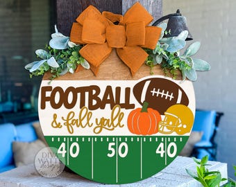May include: A round wooden sign with the words "FOOTBALL & fall y'all" in brown and gold. It features a football, pumpkin, and helmet. The sign is decorated with an orange bow and greenery, and has a football field design.