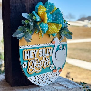 May include: A wooden door hanger shaped like an egg with a yellow, blue, and white polka dot bow, green and yellow background, and the text "Hey Silly Goose".