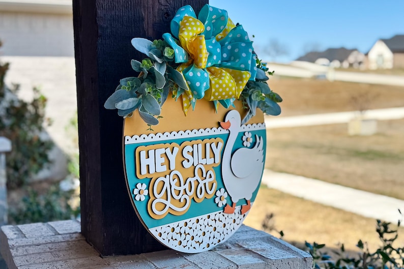May include: A wooden door hanger shaped like an egg with a yellow, blue, and white polka dot bow, green and yellow background, and the text "Hey Silly Goose".