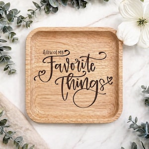 Può includere: Un vassoio quadrato in legno con angoli arrotondati, con la scritta "A Few of My Favorite Things" in elegante corsivo. Il vassoio è circondato da rami di eucalipto e un fiore bianco, creando un'estetica decorativa naturale.