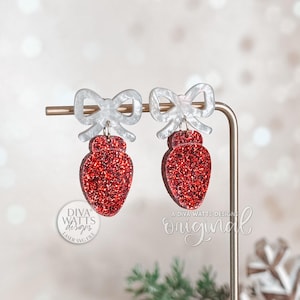 May include: A pair of Christmas light bulb earrings with a red glitter finish. Each earring features a white bow at the top. The earrings are displayed on a gold-colored stand. The Diva Watts Designs logo is visible.