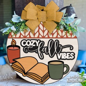 May include: Round wooden sign with the text "Cozy Fall Vibes", a candle, open book, and coffee mug. The sign is decorated with a bow, greenery, and a bell. The sign is perfect for fall decor.