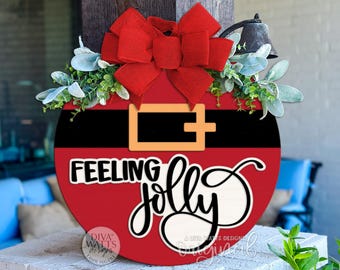 May include: A round, red wooden sign with a black belt design and a gold buckle. The sign has the words "Feeling Jolly" in a whimsical font. A large red bow and greenery adorn the top. A small bell hangs above.