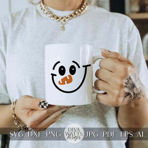 Peut inclure: Une tasse en c&eacute;ramique blanche avec un motif de visage de bonhomme de neige noir et orange. La tasse est tenue par une personne avec un tatouage sur la main.