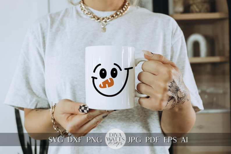 Peut inclure: Une tasse en c&eacute;ramique blanche avec un motif de visage de bonhomme de neige noir et orange. La tasse est tenue par une personne avec un tatouage sur la main.