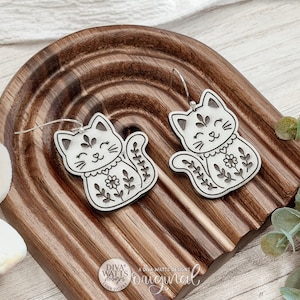 Pode incluir: Um par de brincos pendentes de acrílico branco em forma de gato com detalhes florais e de coração gravados em preto. Cada brinco de gato possui um gancho de metal prateado para pendurar. O design inclui uma pequena flor no peito.
