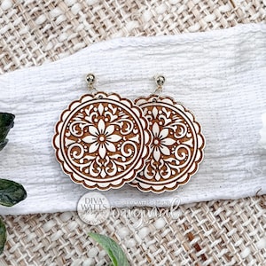 May include: A pair of wooden earrings with a floral design. The earrings are round with a scalloped edge and a detailed, symmetrical pattern. They are light brown with a silver-colored post.