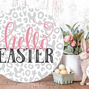 Op de afbeelding: Een rond houten bord met een grijze en witte luipaardprint achtergrond. De woorden "hello easter" zijn geschreven in roze en zwarte letters. Het bord staat op een houten tafel met een klein mandje met eieren en een houten konijnenbeeldje.