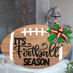 May include: Wooden football-shaped sign with the text "IT'S Football SEASON" in black script. The sign has white stripes and a decorative bow with football and green ribbon accents. A bell hangs above.