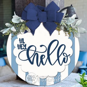 May include: A round wooden door sign featuring vertical light blue and white stripes. The centre has a white scalloped overlay with the dark blue text "HI. HEY. hello". A large dark blue fabric bow and green foliage are attached to the top.