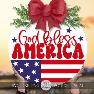May include: A round, white sign with a red bow and green leaves at the top. The sign says "God bless AMERICA" in red letters with a blue and white American flag at the bottom.