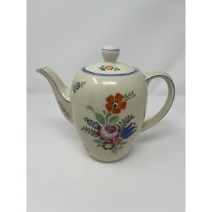 May include: Cream-coloured ceramic teapot with a floral design. The teapot has a rounded body, a curved spout, and a handle. The lid is topped with a small knob. The floral design includes orange, red, blue, and purple flowers with green leaves.
