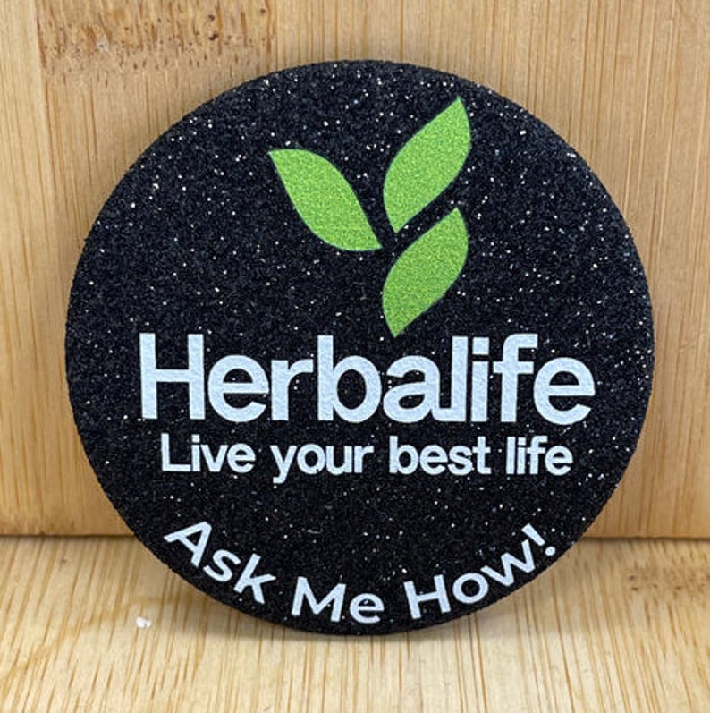 以下が含まれることがあります： 黒と緑のグリッターボタンに「Herbalife 最高の人生を生きよう 聞いてください！」というテキストが書かれています