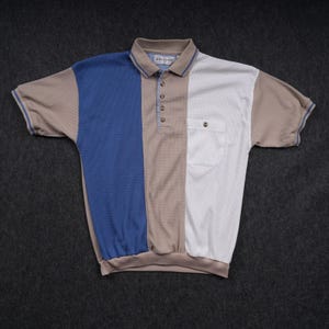 May include: A short-sleeve polo shirt featuring a color-blocked design with a blue left panel, a tan center panel, and a white right panel. The shirt has a collar, a button-up placket, a chest pocket, and ribbed sleeve cuffs with blue trim.