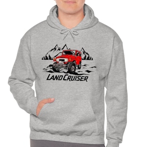 May include: A light grey hoodie with a red Land Cruiser graphic and a mountain scene. The text "LAND CRUISER" is in black. The hoodie has a front pocket and a drawstring hood, typical of casual wear.