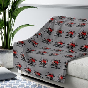 May include: A gray blanket with a repeating pattern of red off-road vehicles and the text "LAND CRUISER". The design includes mountains and a rugged terrain, creating a dynamic and adventurous theme. The blanket is draped over a white sofa.