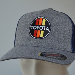 Puede incluir: Una gorra de camionero gris y azul marino con un logotipo de Toyota bordado en negro y rojo. El logotipo presenta la palabra "TOYOTA" en letras blancas y un diseño de rayas rojas, naranjas y amarillas.