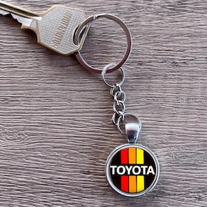 Könnte beinhalten: Ein silberner Schlüssel und ein Schlüsselanhänger mit einem runden Anhänger mit dem Toyota-Logo in weißer Schrift, mit roten, orangefarbenen und gelben Streifen auf schwarzem Hintergrund. Der Schlüssel und der Schlüsselanhänger liegen auf einer Holzoberfläche.