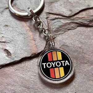 Könnte beinhalten: Ein klarer Acryl-Schlüsselanhänger mit einem silbernen Metallring und einer Kette. Der Schlüsselanhänger hat ein rundes Emblem mit dem Wort "TOYOTA" in weißer Schrift auf schwarzem Hintergrund mit vertikalen Streifen in Rot, Orange und Gelb.