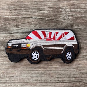 May include: Embroidered patch featuring a brown and white Toyota Land Cruiser. The vehicle has a red and white design on the roof, with the Toyota logo visible. The patch is on a wooden surface.