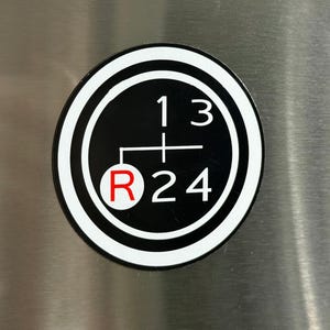 May include: A black and white sticker featuring a gear shift pattern. The sticker has a circular design with the numbers 1, 2, 3, and 4, and the letter R for reverse. The numbers and letter are white, and the R is red.