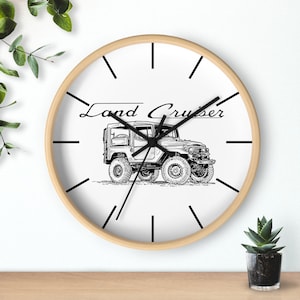 May include: A white wall clock with a wooden frame and black hands. The clock face features a black and white illustration of a Land Cruiser and the text "Land Cruiser".