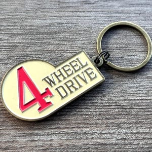 Op de afbeelding: Een gouden sleutelhanger met een wit en rode 4 Wheel Drive-hanger. De hanger is in de vorm van een afgeronde rechthoek met een groot rood cijfer 4 en de woorden "4 Wheel Drive" in zwart.