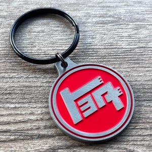Op de afbeelding: Een rode en zilveren sleutelhanger met een zilveren Toyota-logo. Het logo is een gestileerde "T" met een kleine "O" aan de rechterkant.