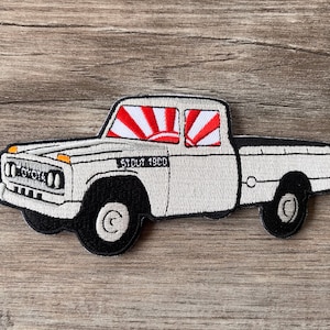 Könnte beinhalten: Gestickter Aufnäher eines weißen Toyota Stout 1900 Pick-ups. Der Truck hat schwarze Räder, eine schwarze Ladefläche und ein Design mit aufgehender Sonne in den Fenstern. Das Toyota-Logo ist vorne sichtbar.