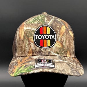 May include: A brown and green camouflage trucker hat with a black and white embroidered Toyota logo in a red, orange, and yellow circle. The hat is adjustable and has a mesh back.