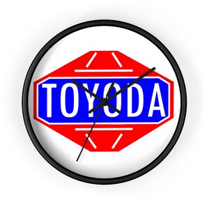 Puede incluir: Reloj de pared redondo con marco negro. La esfera del reloj muestra la palabra "TOYODA" en letras blancas sobre un fondo azul, dentro de un diseño geométrico rojo y blanco. Las manecillas del reloj son negras.