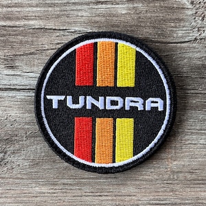 Puede incluir: Un parche bordado en blanco y negro con la palabra "Tundra" en letras blancas. El parche tiene tres rayas verticales en rojo, naranja y amarillo.