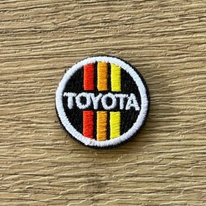 Puede incluir: Un parche bordado circular con fondo negro y tres franjas verticales en rojo, naranja y amarillo. La palabra TOYOTA está bordada en letras mayúsculas blancas en el centro, rodeada por un borde circular grueso de color blanco.
