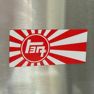 May include: Rectangular sticker featuring a red and white design reminiscent of the Japanese flag. The sticker has a red circle with white Japanese characters inside, surrounded by red and white rays.
