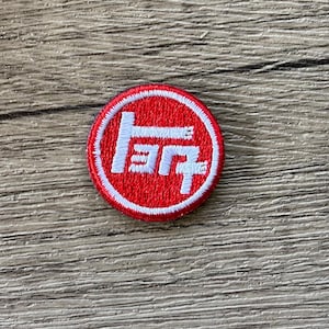Puede incluir: Un parche bordado circular que presenta un fondo rojo con un borde blanco y un logotipo de carácter japonés estilizado en blanco en el centro. El parche tiene una apariencia texturizada y cosida con un patrón de hilo consistente.