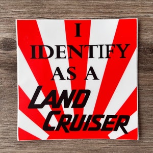 May include: A red and white sticker with a rising sun design. The sticker says "I identify as a Land Cruiser".