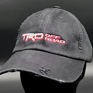 May include: Black baseball cap with a distressed look. The cap has red and white embroidered text that reads "TRD OFF ROAD".