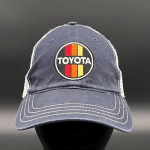 May include: A navy blue trucker hat with a white mesh back. The hat features a vintage-style Toyota logo with red, orange, and yellow stripes.