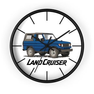 May include: A round wall clock with a black frame and white face. The clock features a blue Land Cruiser illustration with the text "LAND CRUISER" in black. Black clock hands indicate the time.