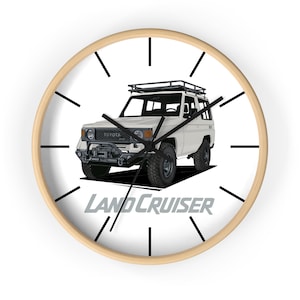 May include: A round wall clock with a light wood frame. The clock face features a white SUV illustration with the text "LAND CRUISER" in gray. Black hands and hour markers complete the design.