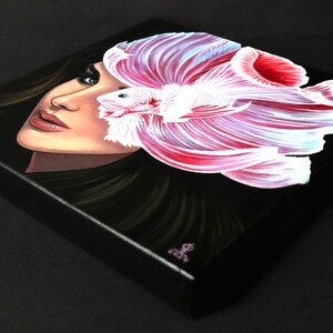 May include: A square canvas painting on a black background featuring a portrait of a person with dark hair and blue eyes. A large, vibrant pink and white betta fish with flowing fins is positioned over the right side of the person's face.