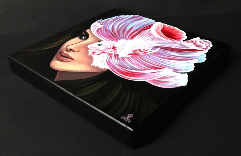 May include: A square canvas painting on a black background featuring a portrait of a person with dark hair and blue eyes. A large, vibrant pink and white betta fish with flowing fins is positioned over the right side of the person's face.