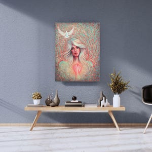 Op de afbeelding: Een canvas kunstafdruk met een vrouw met wit haar en een duif. Het kunstwerk heeft een zachte, etherische kwaliteit met een hartvormige gloed op de borst van de vrouw. De achtergrond is een mix van rode en blauwgroene tinten.