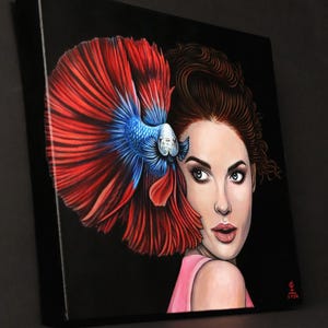 May include: A square canvas painting on a black background featuring a portrait of a person with brown hair and a pink top, positioned next to a large, vibrant betta fish with flowing red and blue fins that frame the side of the person's face.