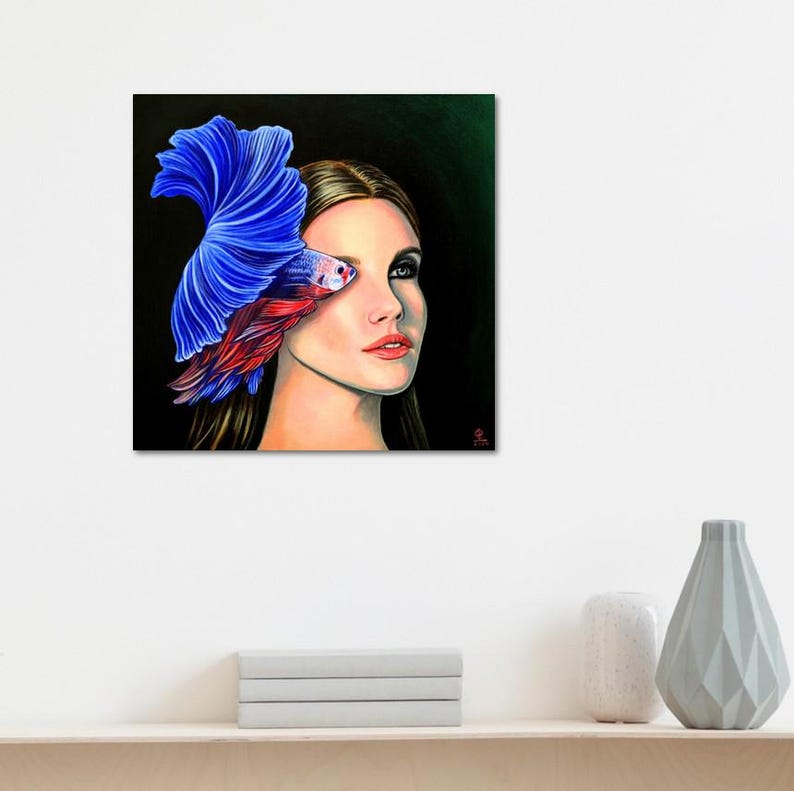 Puede incluir: Una impresi&oacute;n de arte mural cuadrada que muestra el retrato de una persona con cabello casta&ntilde;o largo. Un pez luchador azul y rojo vibrante est&aacute; posicionado sobre el lado izquierdo de la cara, con sus aletas fluidas extendi&eacute;ndose hacia arriba y hacia afuera sobre un fondo oscuro.
