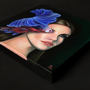 Puede incluir: Un cuadro cuadrado sobre lienzo que muestra el retrato de una persona con cabello largo y oscuro. Un pez luchador de color azul y rojo vibrante se sit&uacute;a sobre el ojo de la persona, con sus aletas extendidas hacia arriba. Fondo negro.