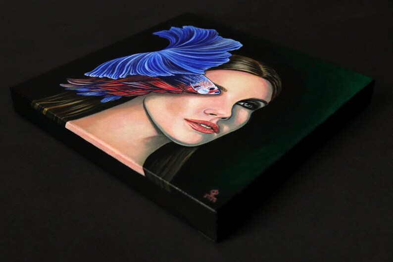 Puede incluir: Un cuadro cuadrado sobre lienzo que muestra el retrato de una persona con cabello largo y oscuro. Un pez luchador de color azul y rojo vibrante se sit&uacute;a sobre el ojo de la persona, con sus aletas extendidas hacia arriba. Fondo negro.
