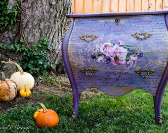May include: A purple and gold painted antique dresser with floral and script details. The dresser has three drawers with ornate brass handles. Several pumpkins of various sizes and colors are in the foreground.